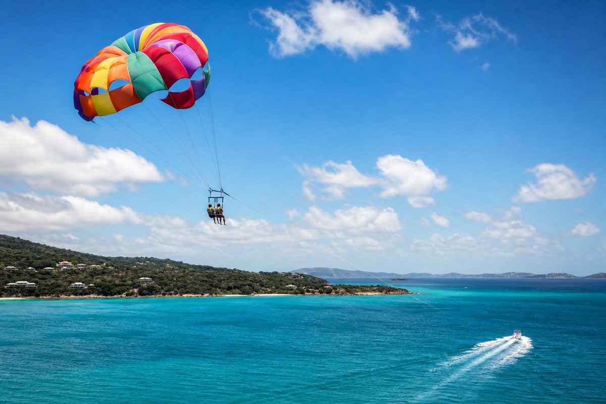 A parasail canopy towed high above turquoise Caribbean water with a reef and white-sand beach below